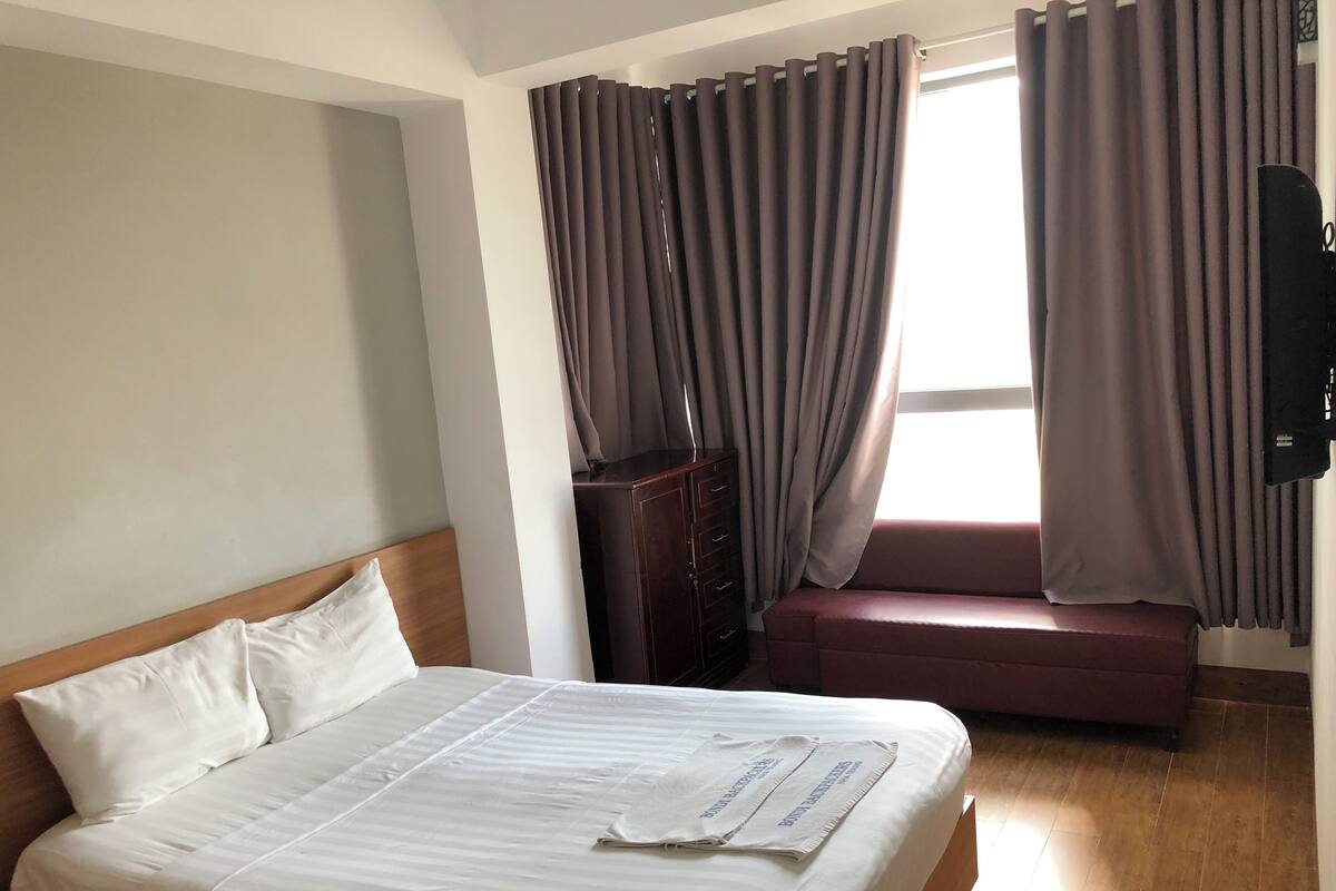 Hotelkamer met een bed, houten hoofdbord, een houten dressoir, een bankje en een raam met gordijnen.