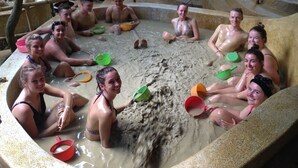 Un groupe de personnes est assis dans un grand bain à remous extérieur, entouré de différents seaux et muni d’une fontaine d’eau.