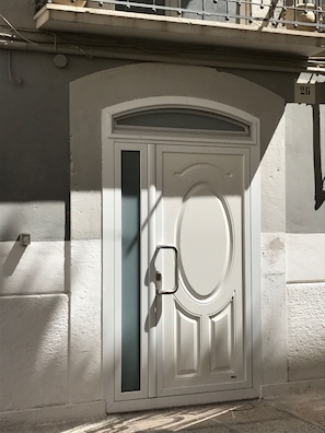 A white door with a glass panel and a handle, set in a building with a textured wall and a balcony above. - B&B Bari Old Town (Bari)