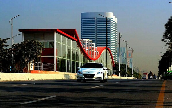 A modern building with a unique red structure in front, a car on the road, and a tall building in the background.