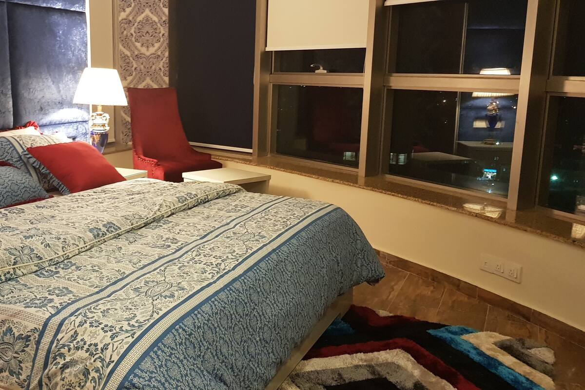 A neatly made bed with patterned bedding, a bedside table with a lamp, and a view of the city through a window.