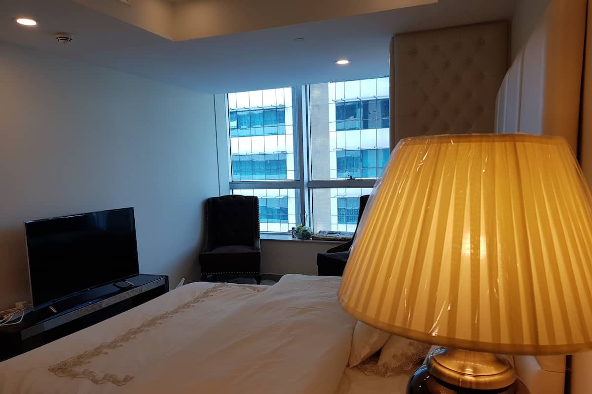 A hotel room with a bed, a TV, a chair, a window with a view of buildings, and a lamp.
