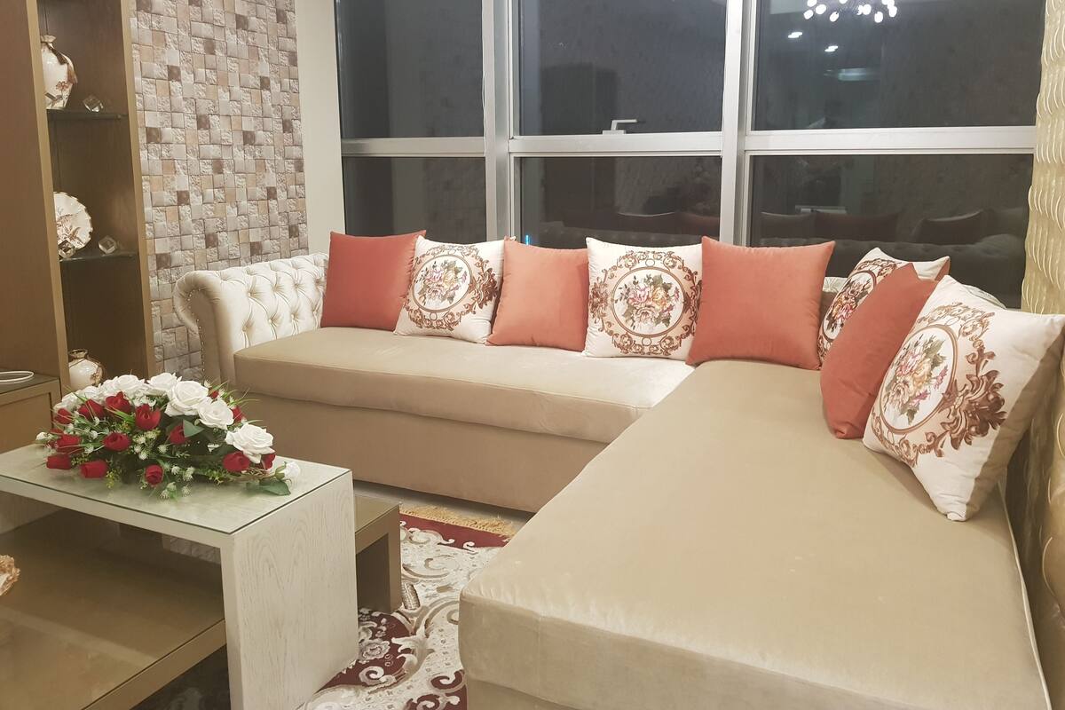 A living room with a beige sectional sofa, a glass coffee table with a floral arrangement, and a brick-patterned wall.