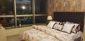 A bedroom with a large window, a bed with floral bedding, a wooden headboard, and a nightstand with a lamp.