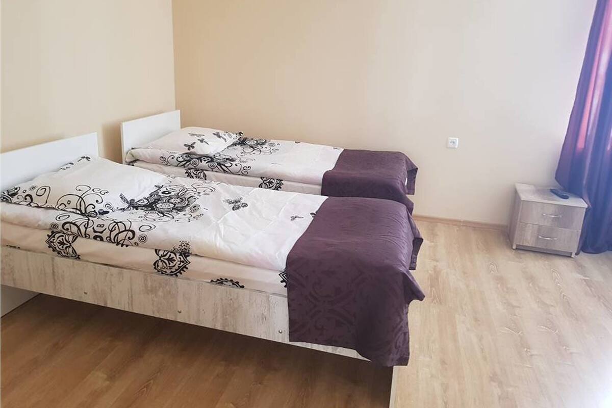 Une chambre avec deux lits simples, chacun recouvert d’une couvre-lit à motifs et d’une couverture violette. On y trouve une petite table de chevet avec une lampe et un tiroir. La chambre est dotée d’un plancher en bois et d’un mur uni.