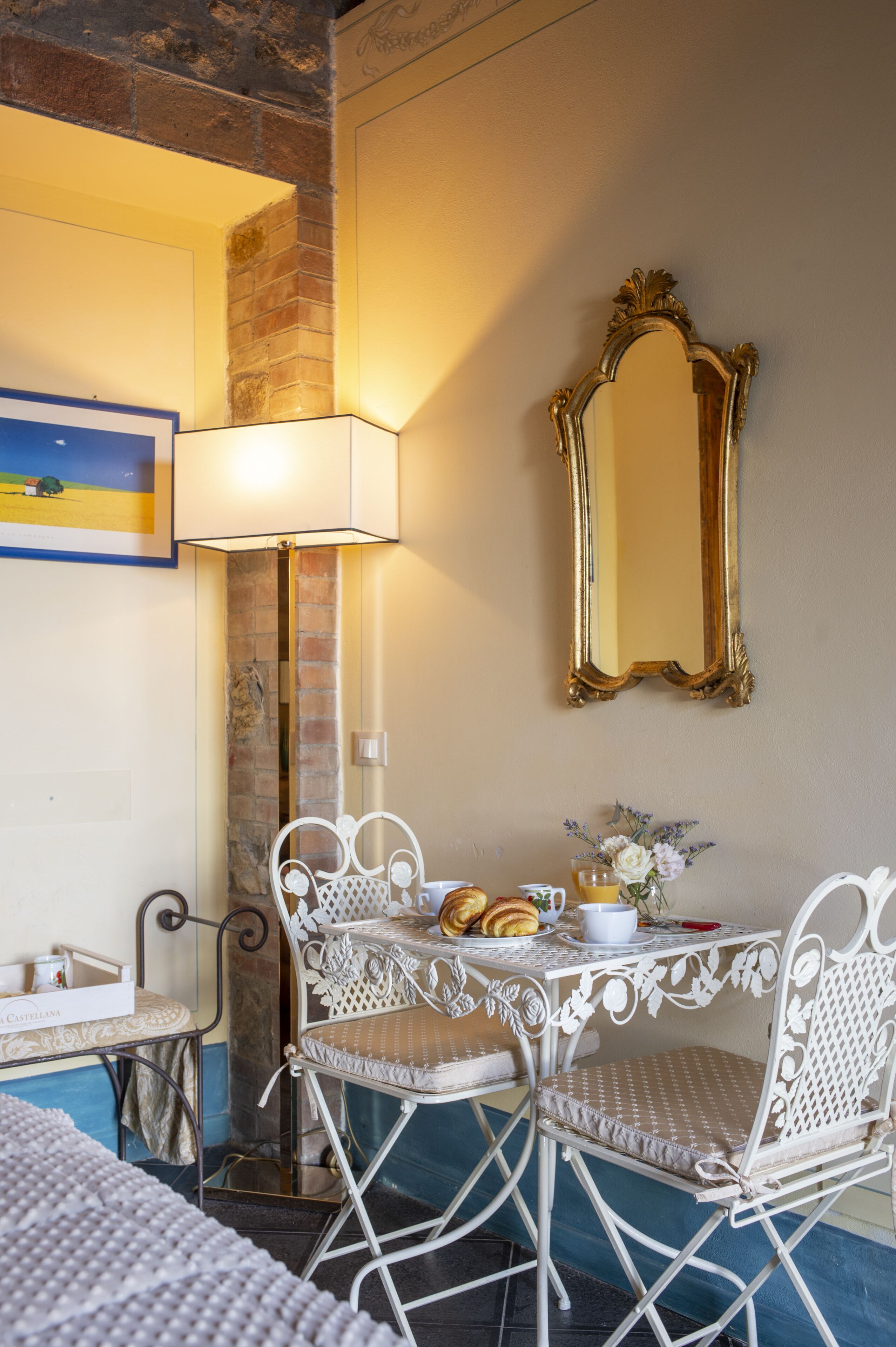 Um quarto com uma mesa e cadeiras de metal branco, uma luminária, um espelho e um quadro na parede.