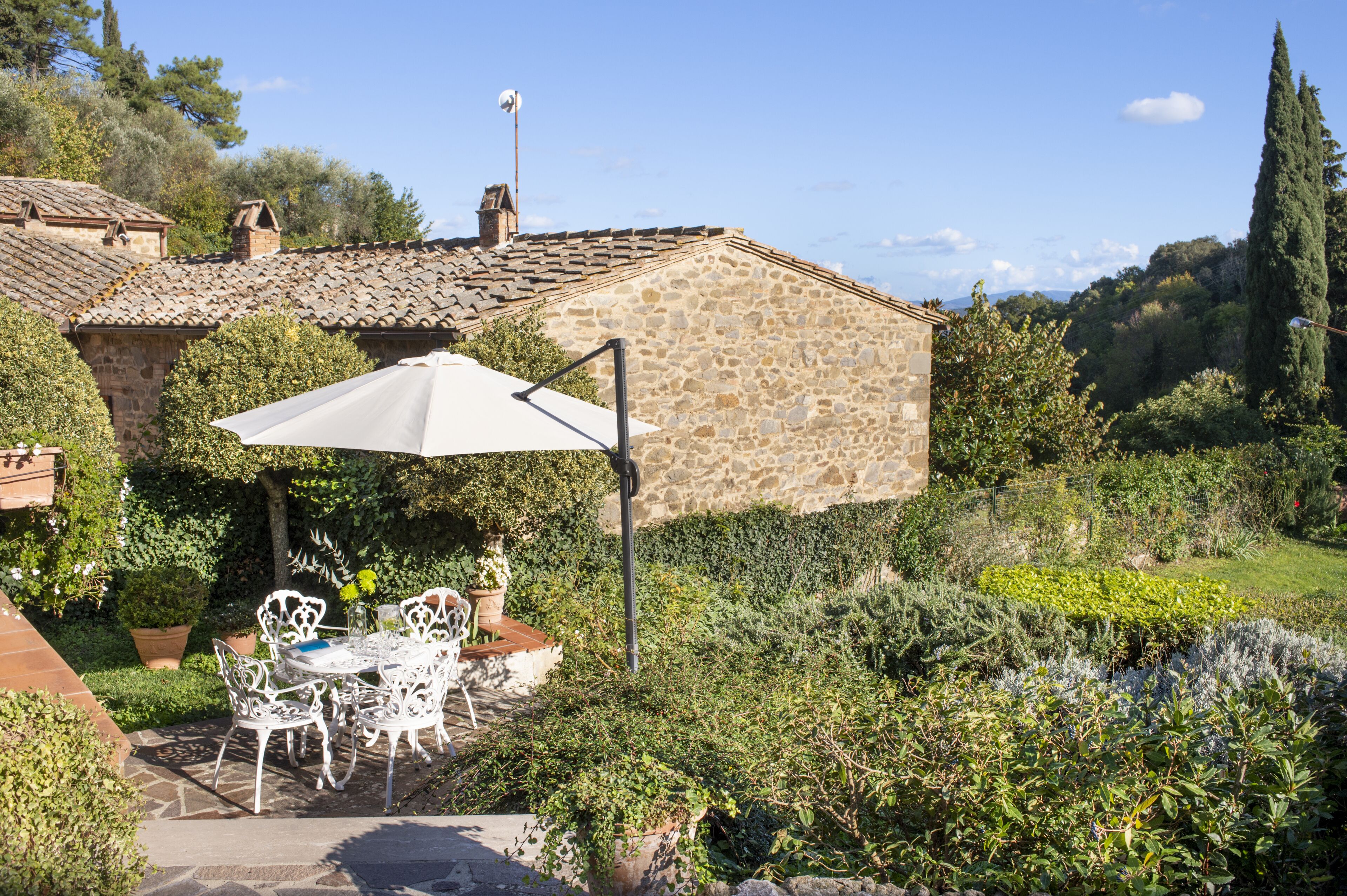 Una casa de piedra con techo de tejas, rodeada de exuberante vegetación y un patio con mobiliario exterior.
