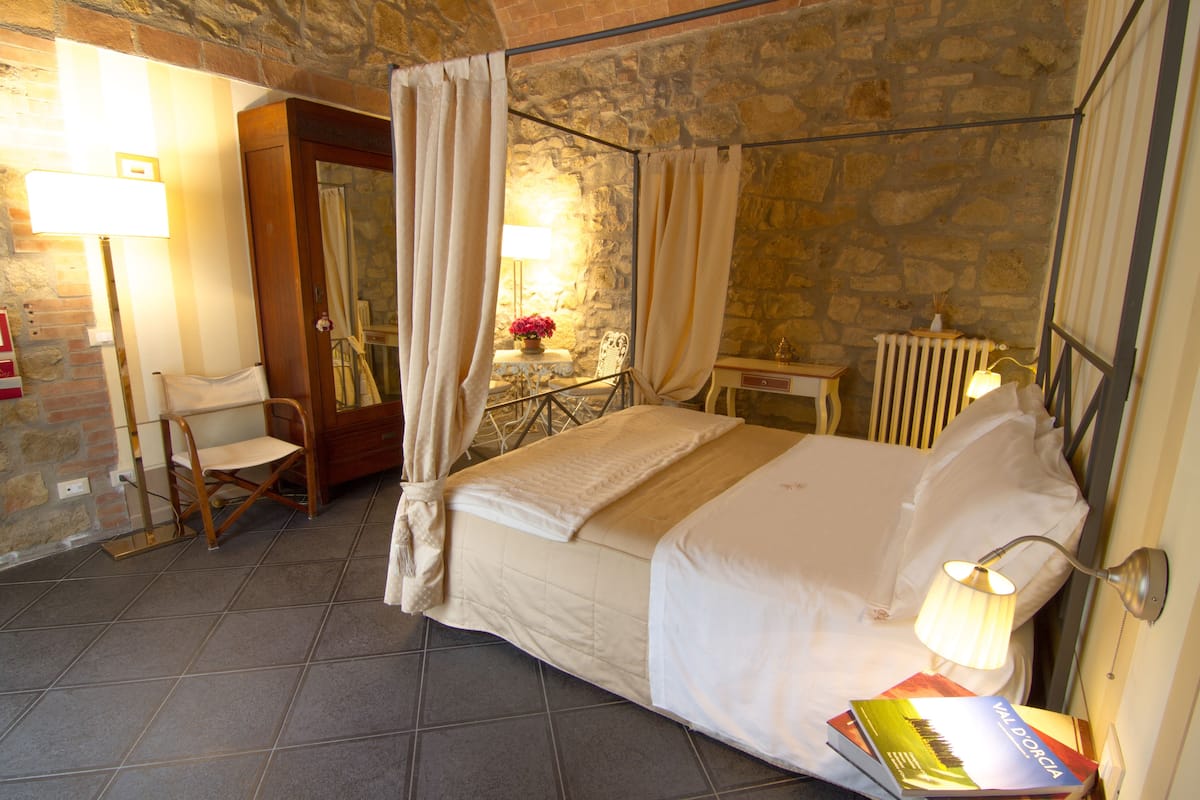 Ein Schlafzimmer mit Bett, einem Sessel, einem Tisch mit Lampe und Blick auf eine Steinmauer.