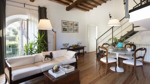 A spacious living room with a white sofa, wooden dining table, and chairs. There is a staircase with a view of the outdoors. - B&B Porta Castellana (Montalcino)