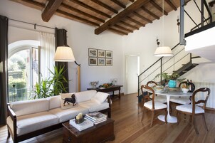 A spacious living room with a white sofa, wooden dining table, and chairs. There is a staircase with a view of the outdoors.