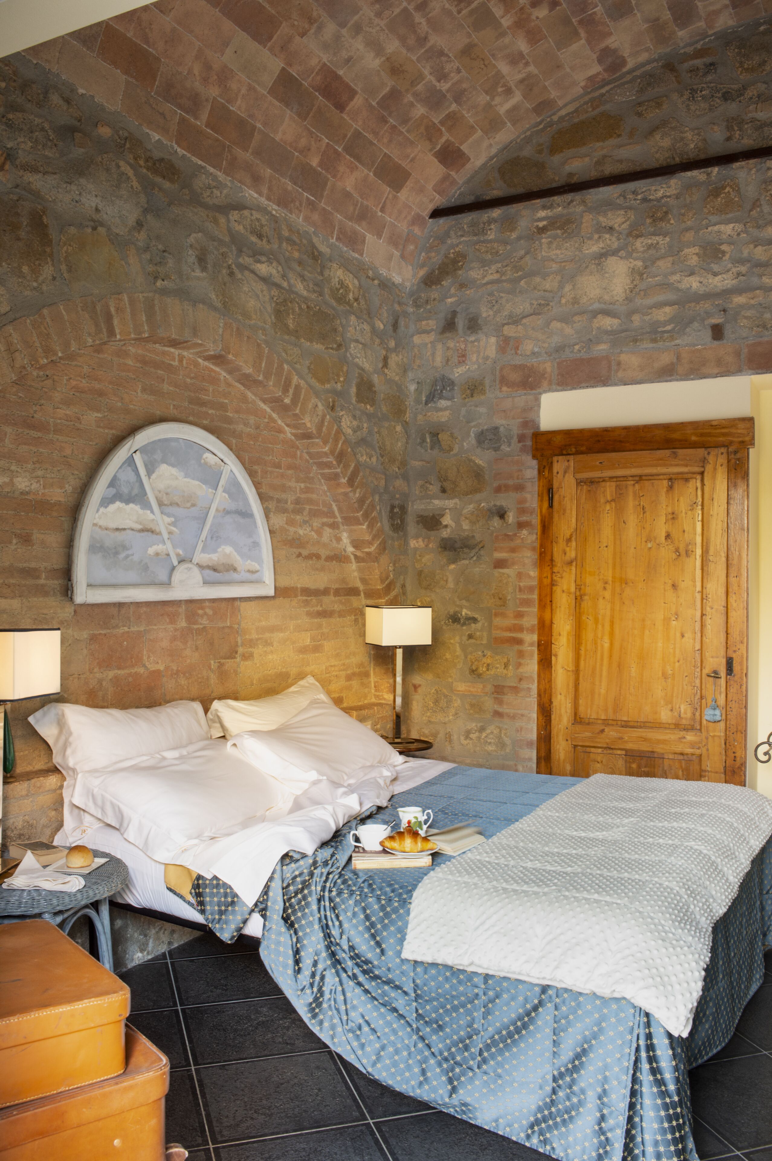 A bedroom with a brick wall, a wooden door, a bed with white bedding, a blue patterned bedspread, and a small table with a lamp and a cup.