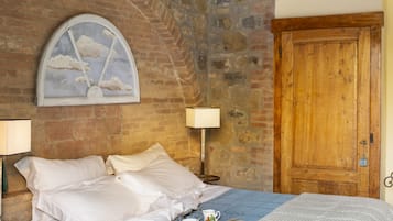 A bedroom with a brick wall, a wooden door, a bed with white bedding, a blue patterned bedspread, and a small table with a lamp and a cup.