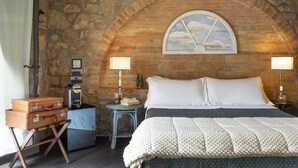A bedroom with a stone wall, a bed with white linens, two bedside lamps, a small table with a lamp, and a suitcase on a stand. - B&B Porta Castellana (Montalcino)