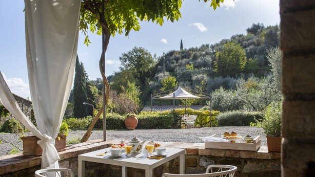 An outdoor dining area with white tables and chairs, a white canopy, and a view of a hillside with trees and a building in the background. - B&B Porta Castellana (Montalcino)