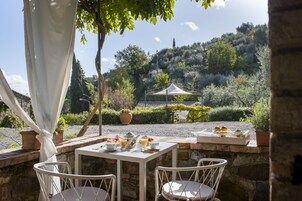 Un área de comedor al aire libre con mesas y sillas blancas, un toldo blanco y vistas a una colina con árboles y un edificio al fondo.