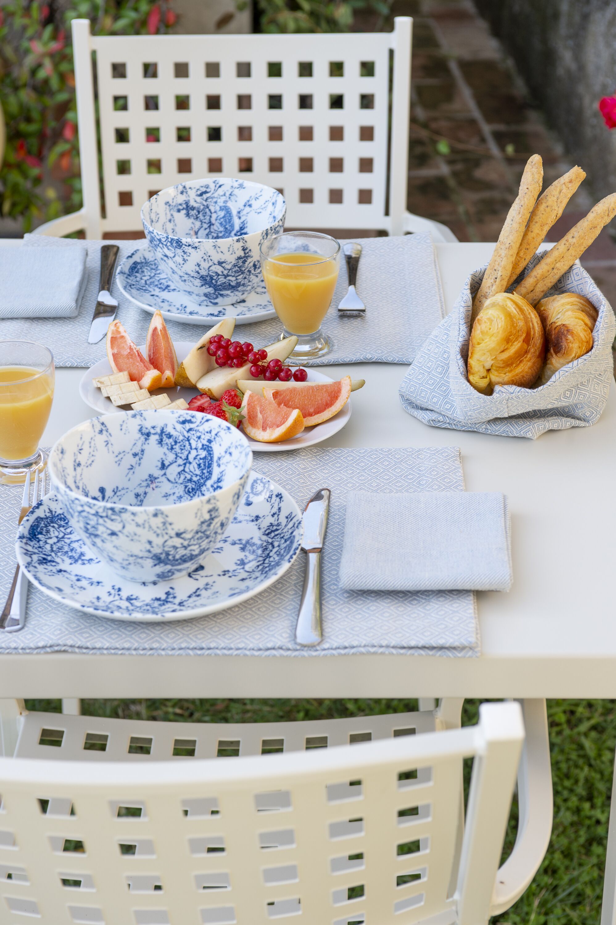 Une table dressée pour le petit-déjeuner, avec des croissants, des fruits et du jus, des chaises blanches et une nappe à motifs.