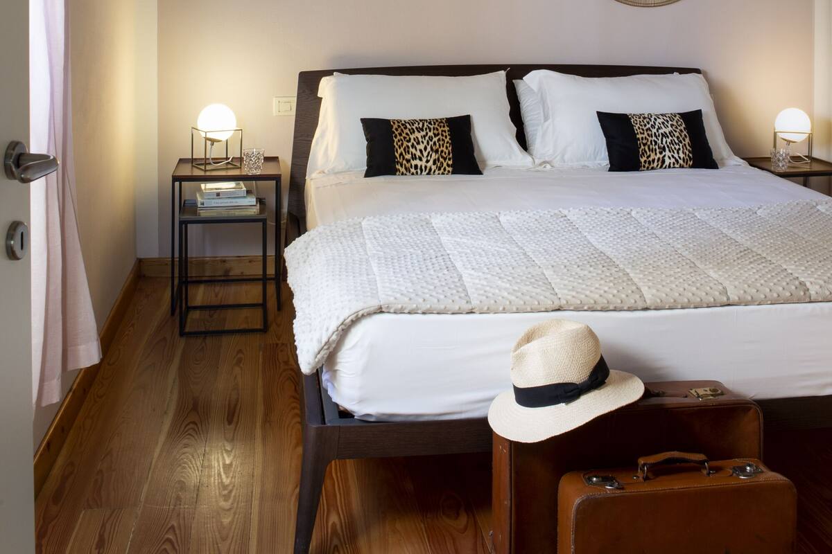 Ein Schlafzimmer mit einem großen Bett, Holzboden, einem runden Spiegel an der Wand und einem Koffer auf dem Boden.