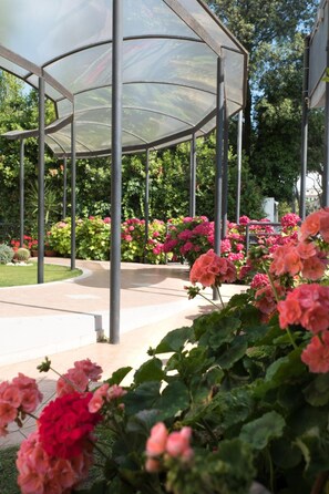 A covered outdoor area with a glass roof, surrounded by greenery and flowering plants. - Hotel Maita (Camaiore)