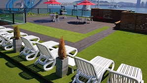 A rooftop terrace with white lounge chairs, a small table, and red umbrellas. A bridge and city skyline are visible in the background. - The Blue Guesthouse - Hostel (Busan)