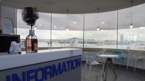 A reception area with a sign reading "INFORMATION," a decorative feather, and a view of a bridge and buildings through large windows. - The Blue Guesthouse - Hostel (Busan)