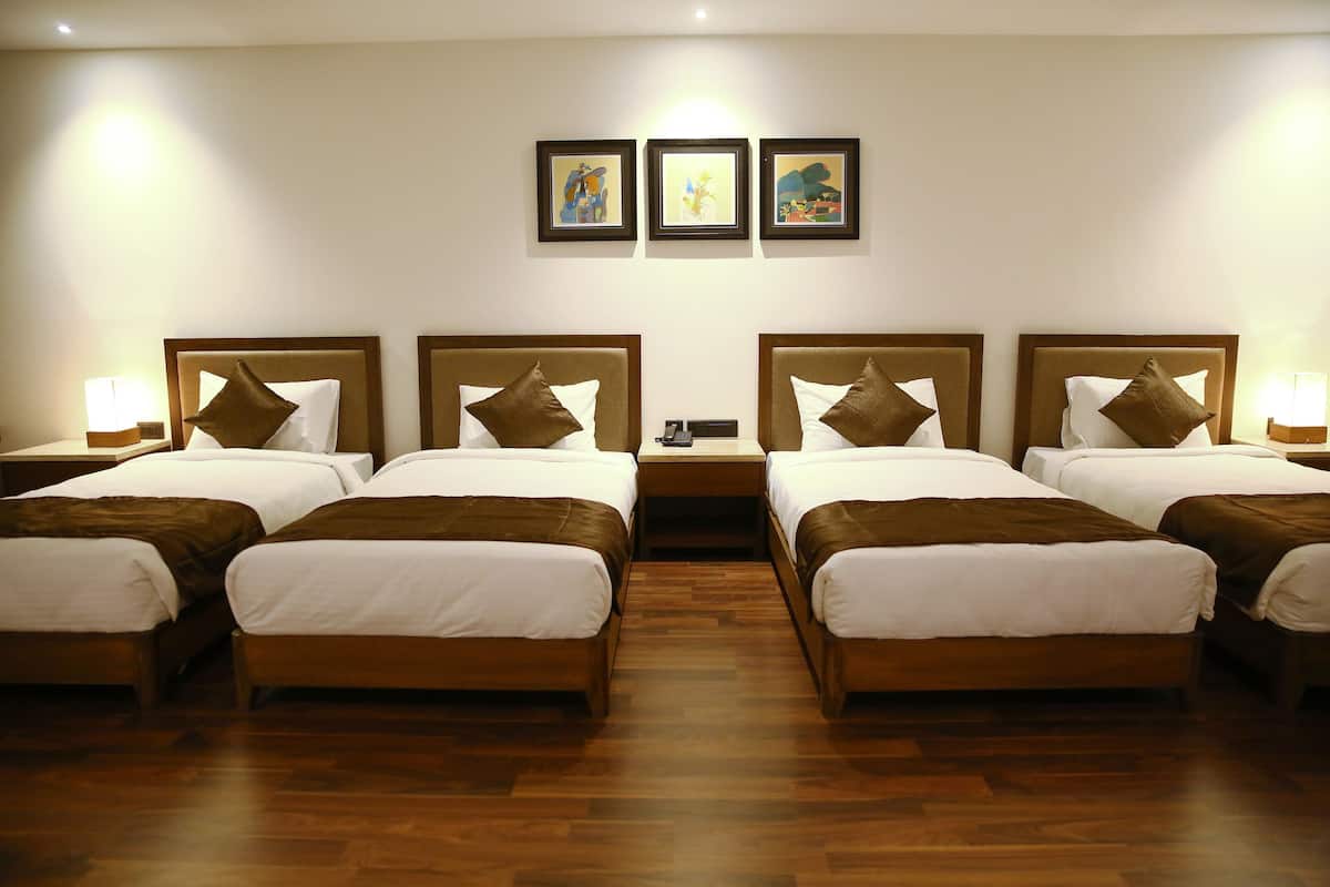 A hotel room with four beds arranged in a row, each with a wooden headboard and white bedding. There are framed artworks on the wall above the beds.