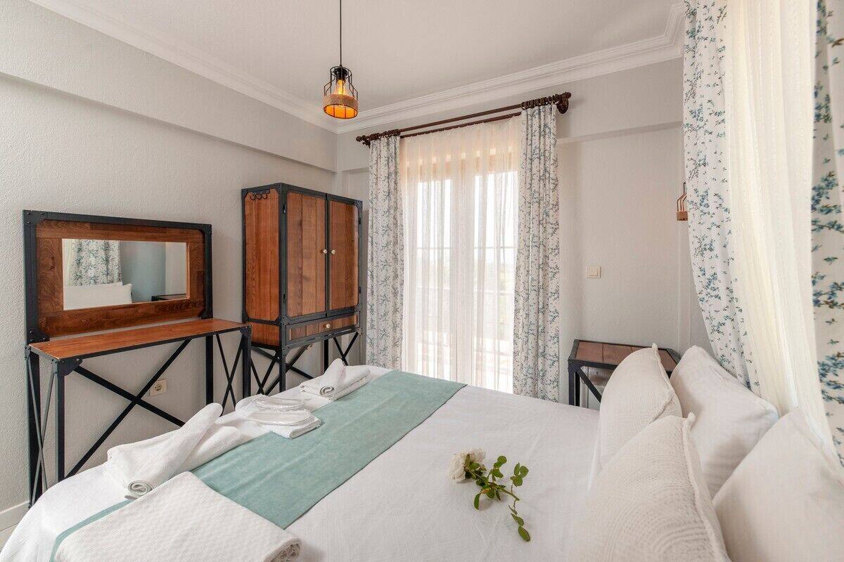 Un lit bien fait, recouvert d’un plaid vert, une armoire en bois, un miroir et une fenêtre ornée de rideaux légers.