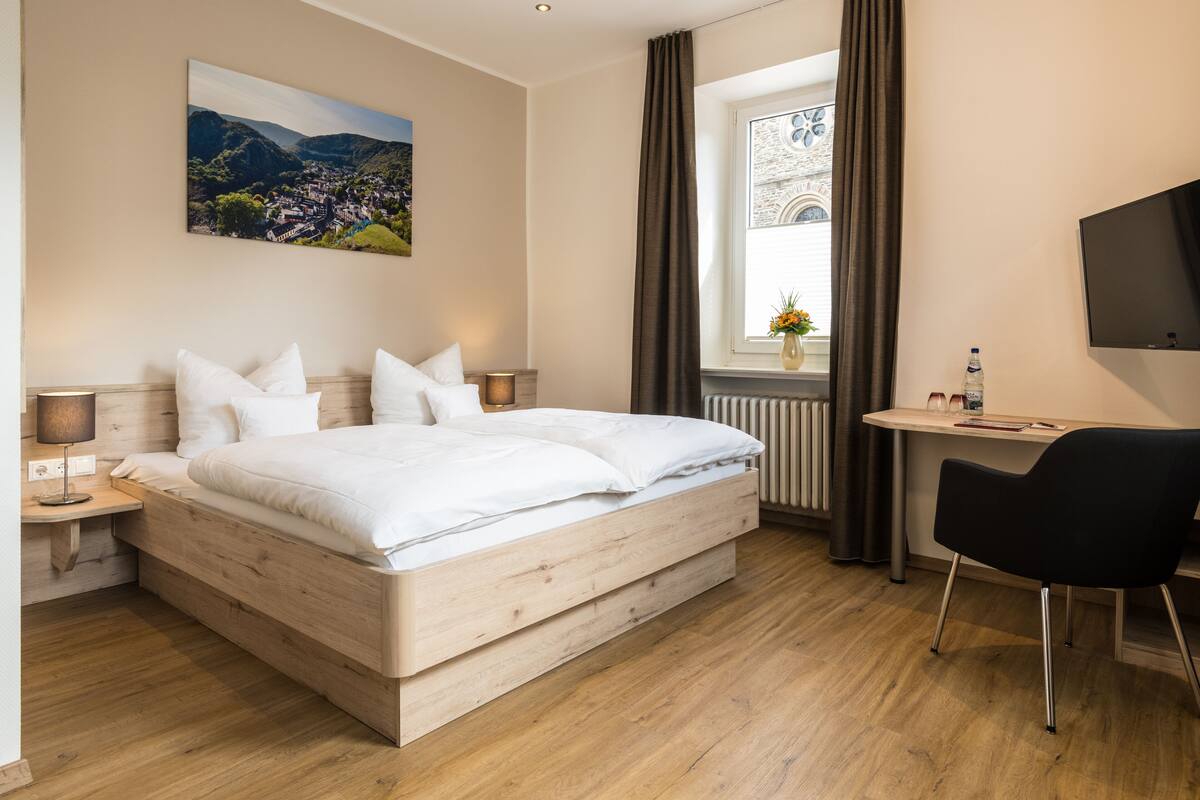 Ein modernes Hotelzimmer mit einem hölzernen Bett, einem Schreibtisch, einem Stuhl, einem Fernseher und einem Fenster mit Vorhängen.