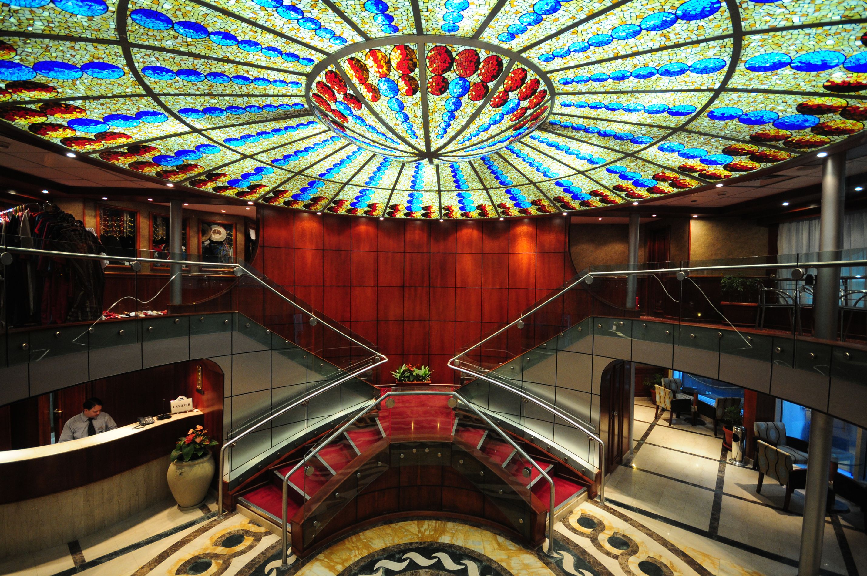 A hotel lobby with a grand staircase, a reception desk, and a ceiling with a stained glass design.