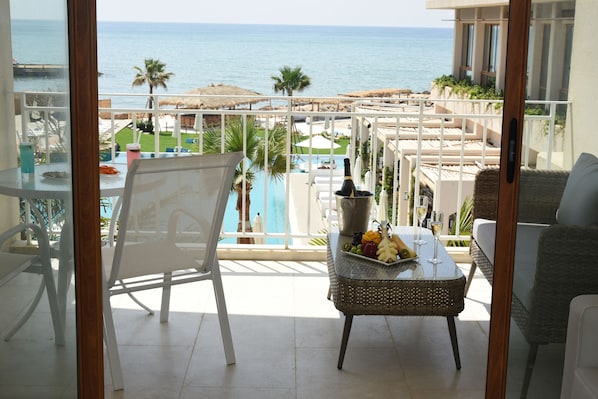 A balcony with a view of the sea, a pool, and a dining area with a table and chairs.