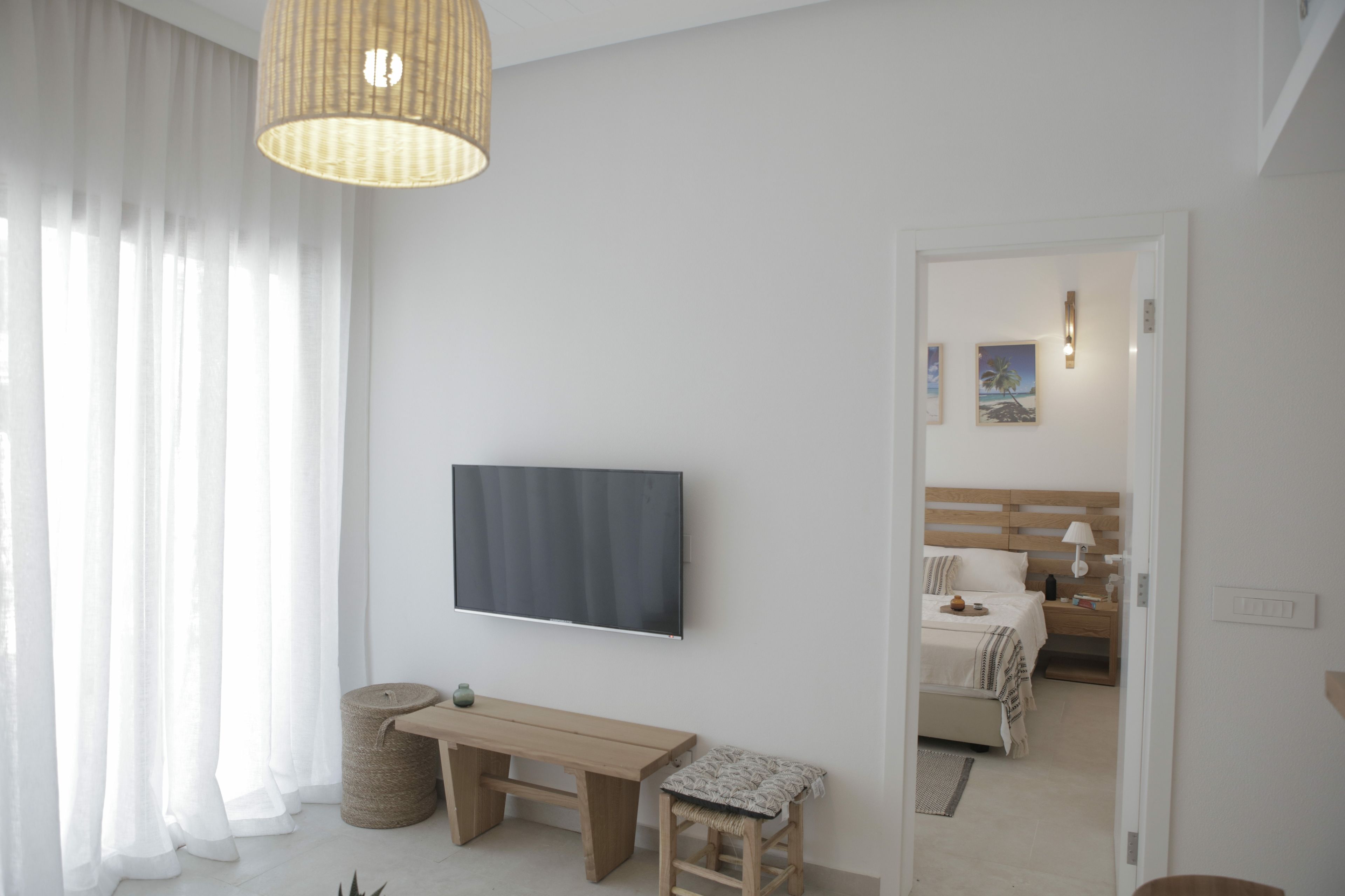 A modern bedroom with a large flat-screen TV mounted on the wall, a wooden desk, a bench, and a bedside table with a lamp.