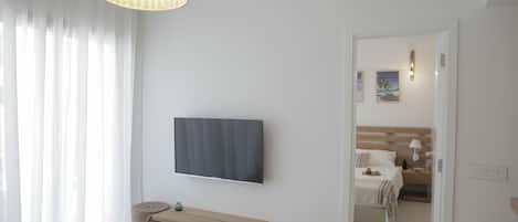 Un dormitorio moderno con una televisión de pantalla plana grande montada en la pared, un escritorio de madera, un banco y una mesita de noche con una lámpara.