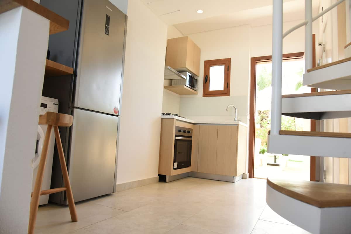 Una cocina moderna con horno empotrado, microondas y refrigerador de acero inoxidable. Hay un taburete de madera y una ventana sobre el fregadero.