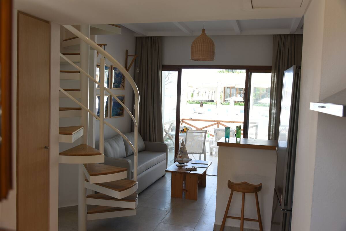 Un interior moderno con una escalera en espiral, un sofá, una mesa de comedor y vistas amplias a través de grandes ventanales.