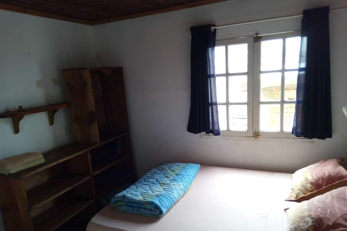 Ein kleiner, schlichter Raum mit einem Bett, einem Fenster mit Vorhängen, einem Holzregal und einem an der Wand befestigten Regal.