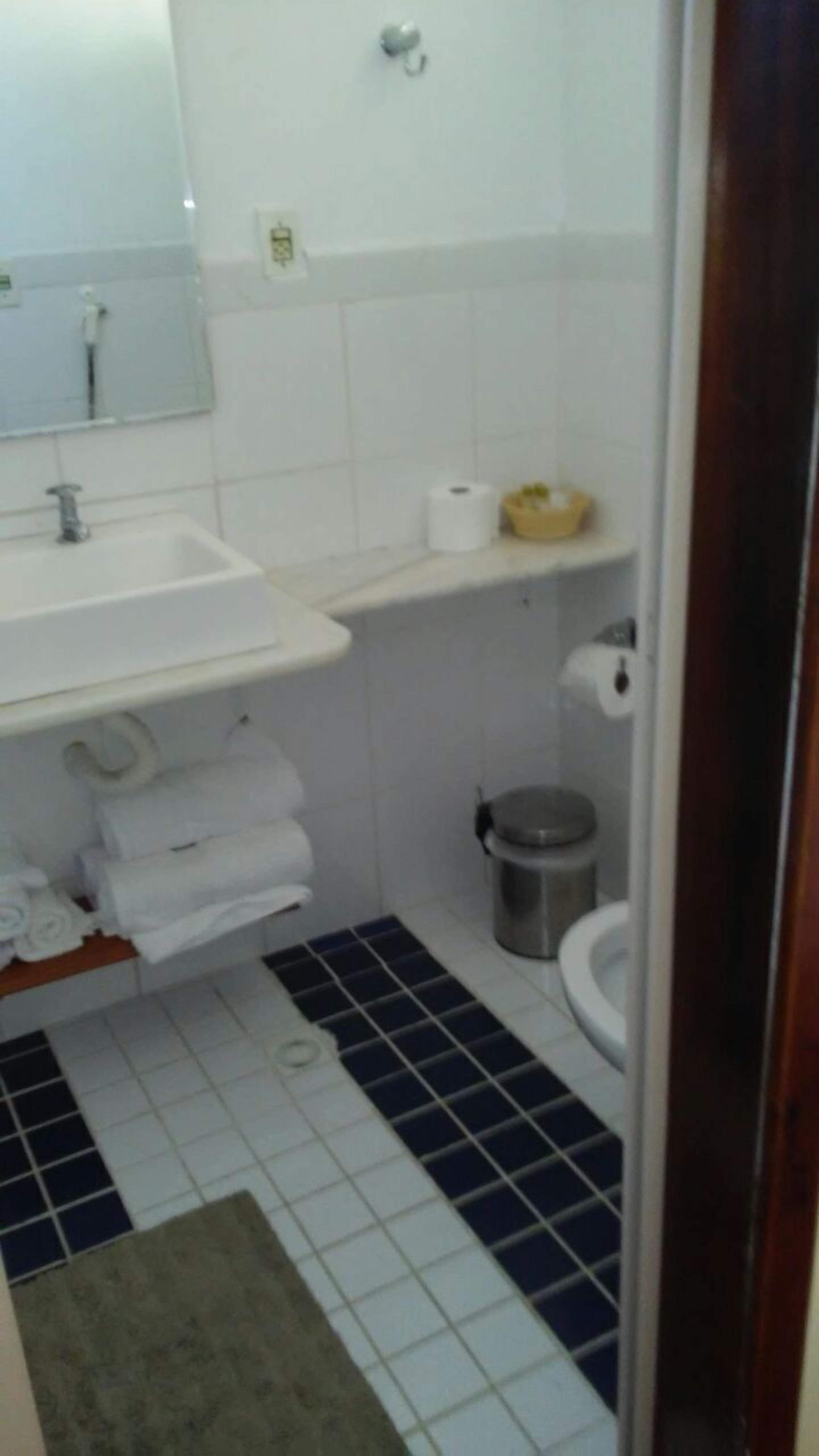 Een badkamer met een witte wastafel, spiegel en toilet. Er is een prullenbak en een klein plankje met toiletartikelen.
