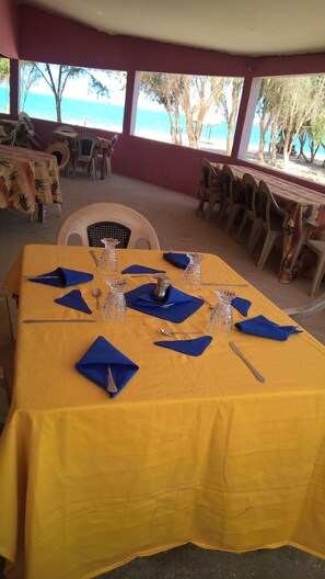 A dining table set with blue napkins and silverware, surrounded by plastic chairs, with a view of the beach and trees through large windows.