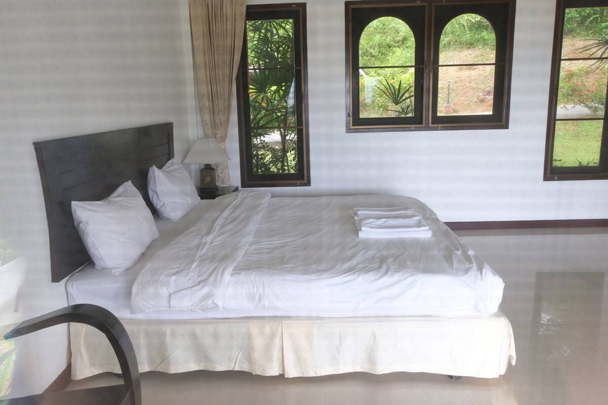 Une chambre à coucher comprenant un lit, une table de chevet, une chaise et de grandes fenêtres donnant sur un paysage verdoyant.
