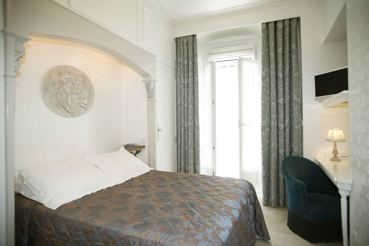 Een slaapkamer met een bed, een bureau, een stoel, een televisie en een deur naar een balkon.