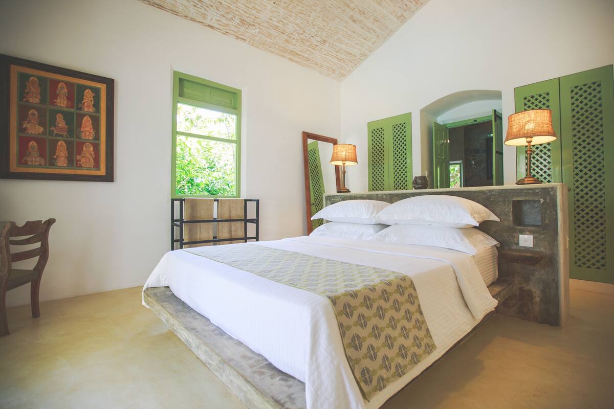 Uma cama bem arrumada com colcha estampada, uma cadeira de madeira, uma janela com persianas verdes e um espelho com moldura verde.