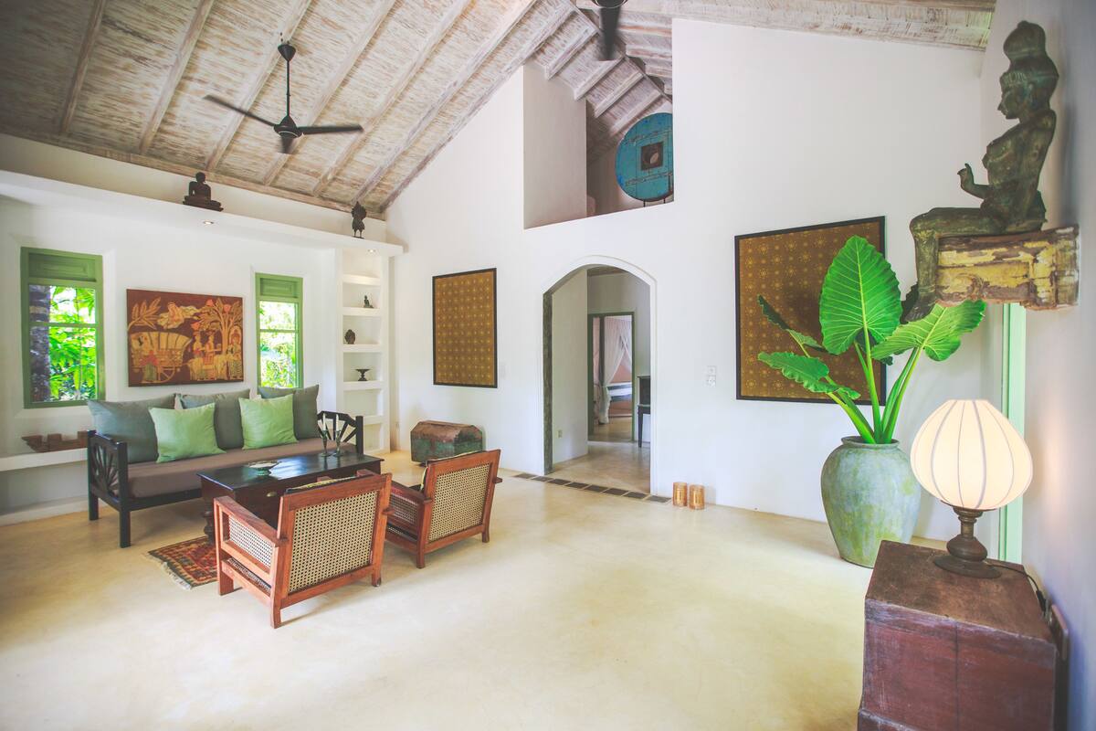 Uma sala de estar espaçosa com teto alto, ventilador de teto, sofá com almofadas verdes, mesa de jantar com cadeiras e uma planta grande.