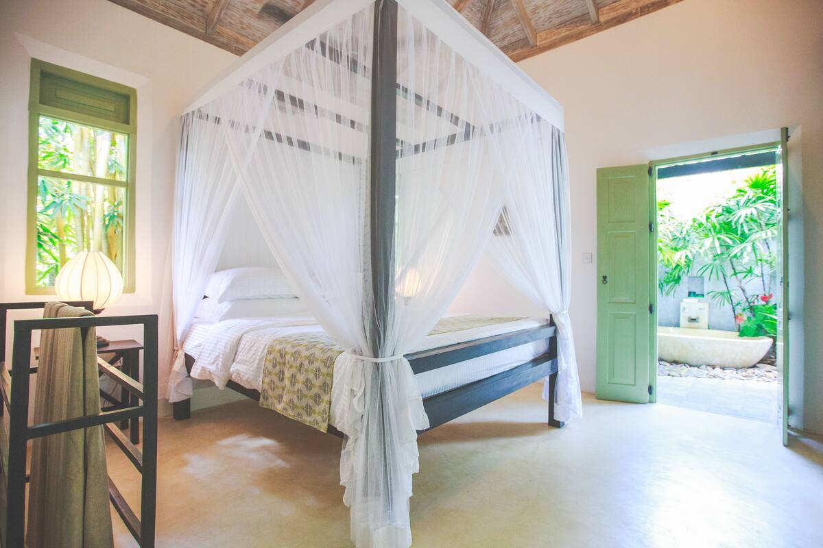 Uma cama com dossel branco, uma porta verde e uma janela com vista para o verde.