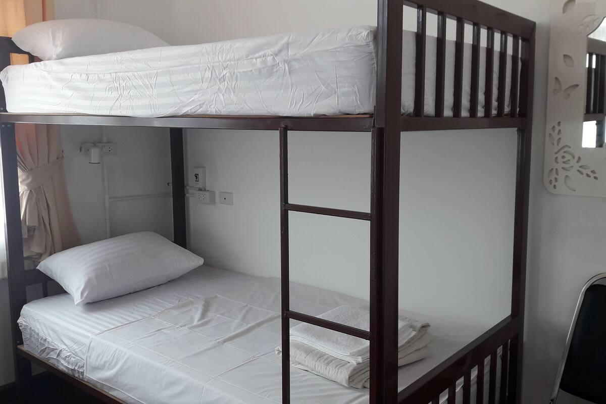 Uma cama beliche com roupa de cama branca, uma mesinha pequena e uma cadeira em um quarto com janela e luminária fixada na parede.