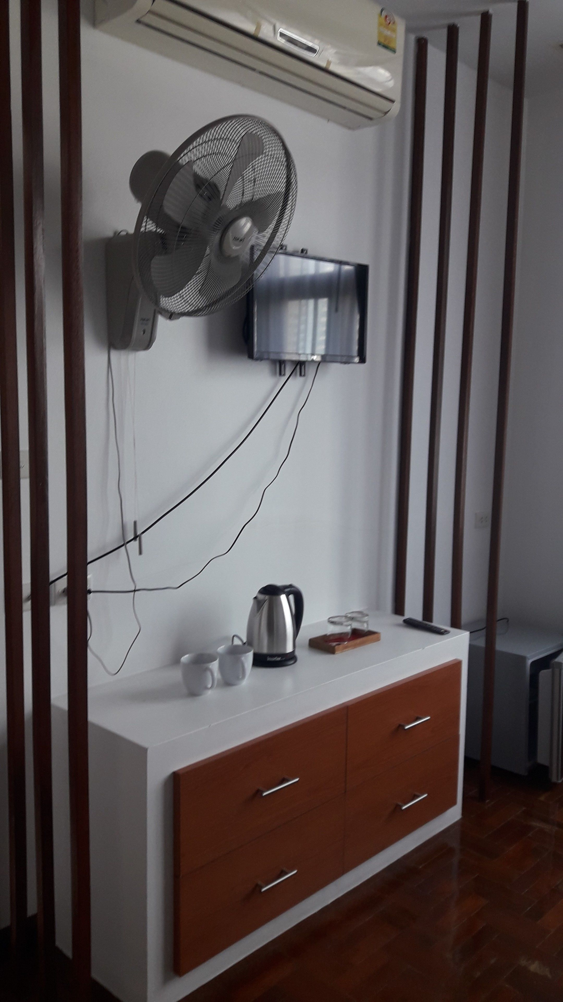 Une pièce avec un climatiseur blanc fixé au mur, un ventilateur mural, une petite télévision, une bouilloire et un meuble en bois avec des tiroirs.