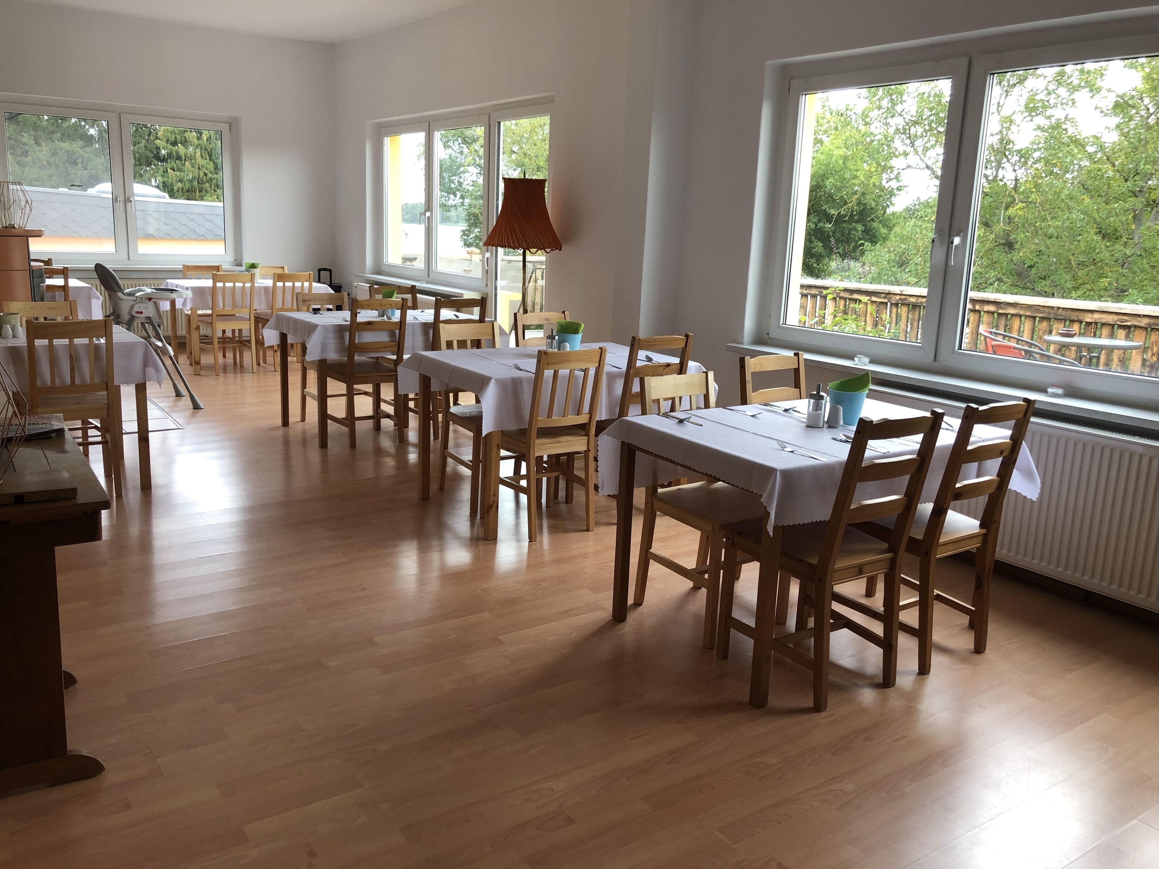 A dining area with multiple tables and chairs, large windows with a view of greenery, and a radiator under the windows.