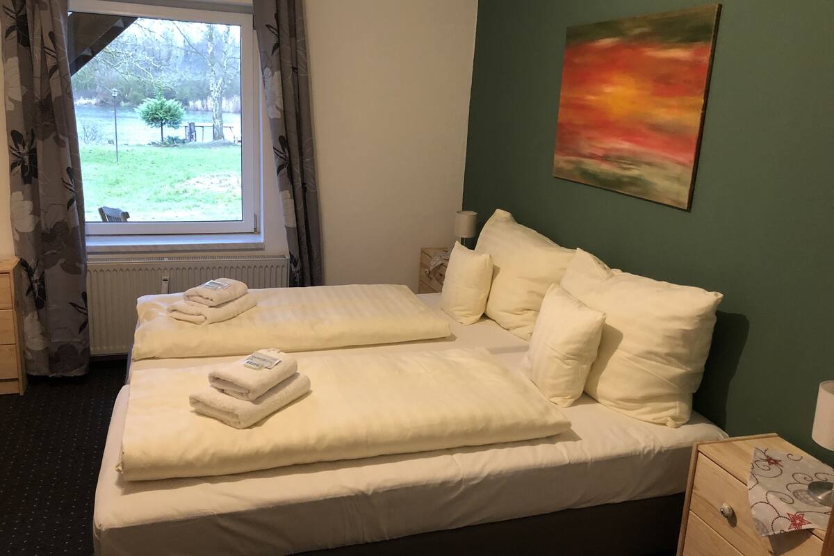 A double bed with white linens and pillows, a radiator, a window with curtains, a painting on the wall, and a bedside table with a lamp.