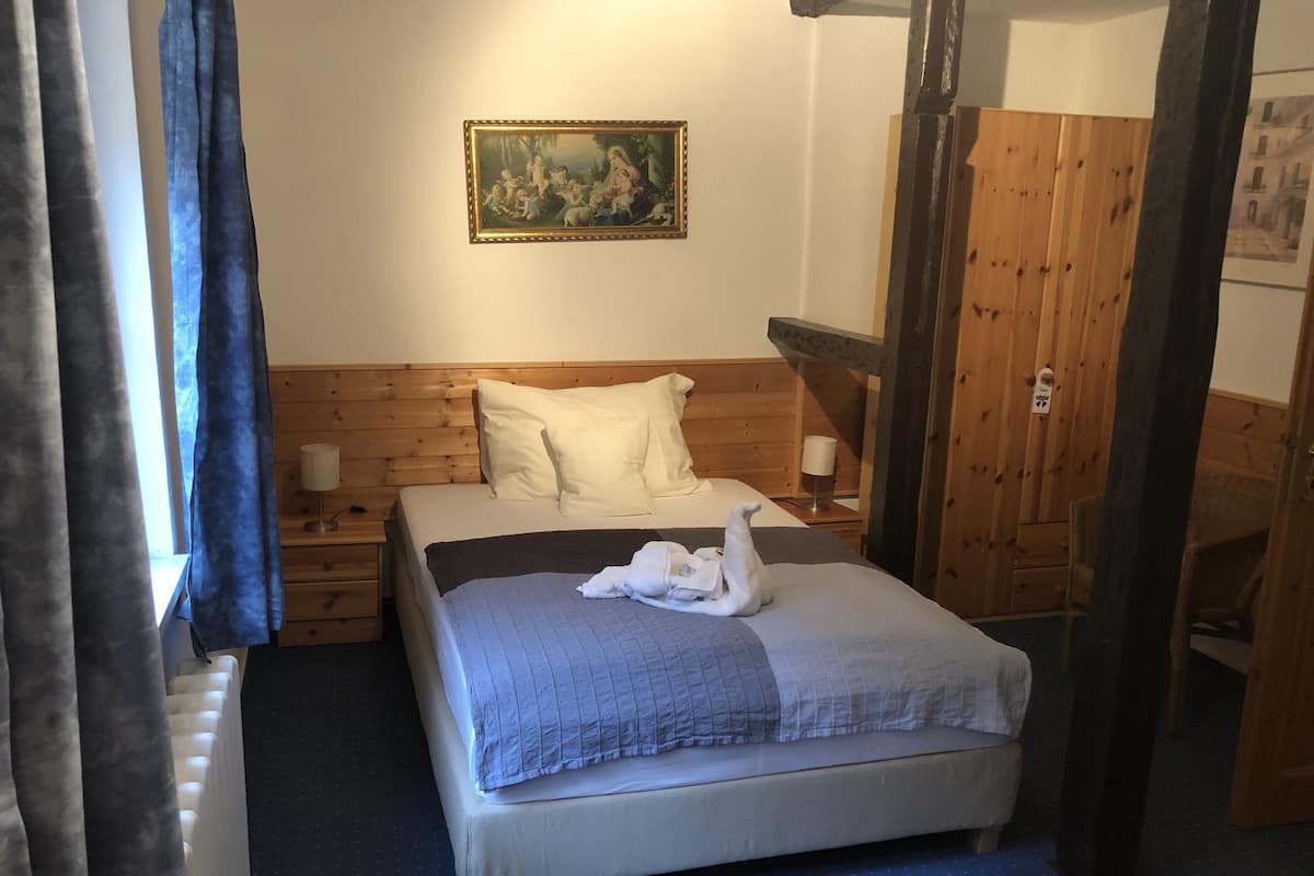 A bedroom with a wooden headboard, a bed with a blue quilt, a white swan-shaped towel, a painting on the wall, and a wooden wardrobe.