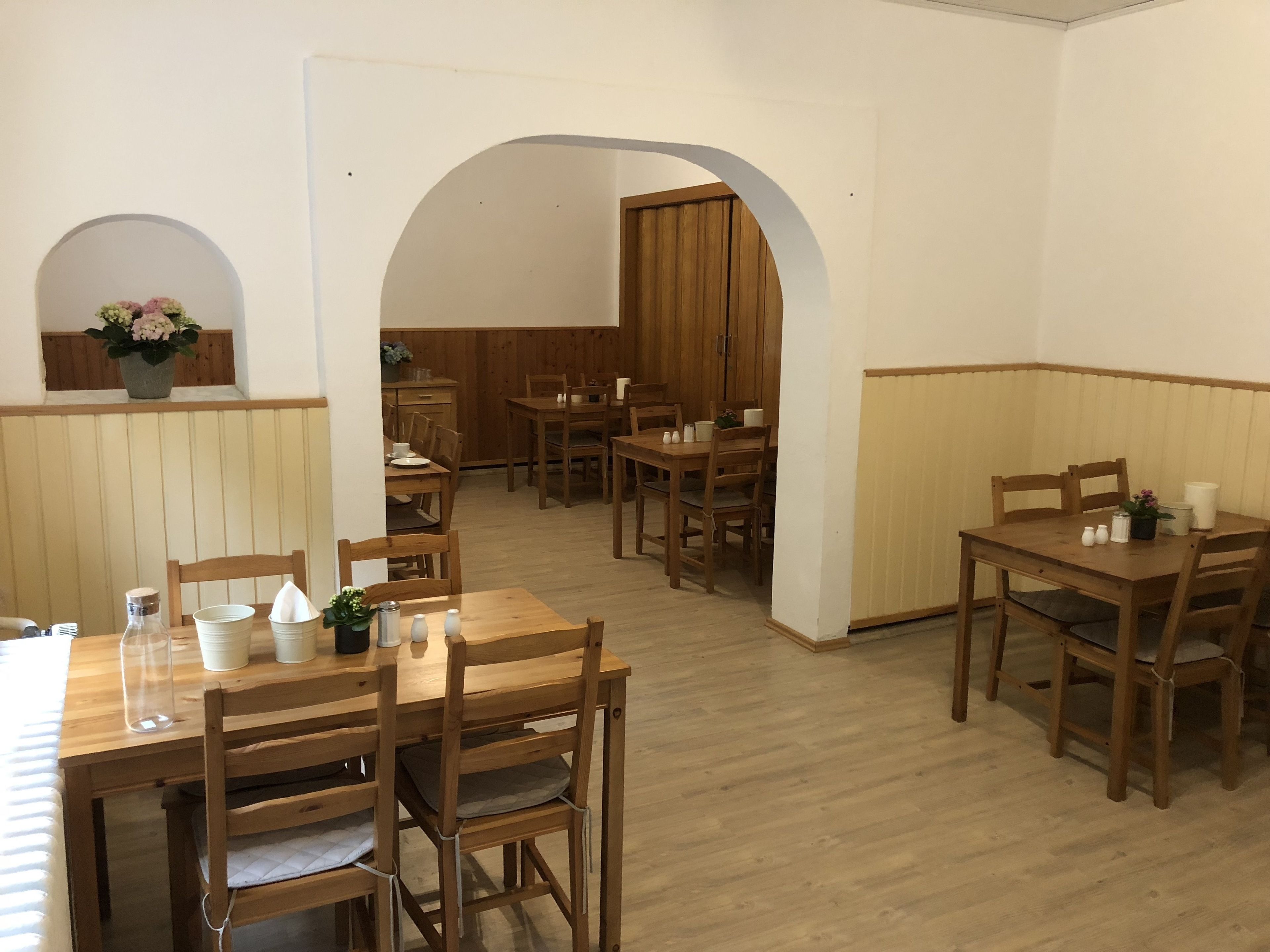 A dining area with wooden tables and chairs, a kitchen in the background, and a small alcove with a plant.