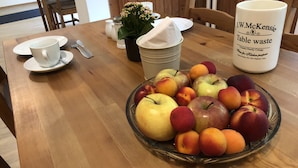 A wooden dining table with a fruit bowl containing apples and peaches, a white mug with text, salt and pepper shakers, and a small potted plant.