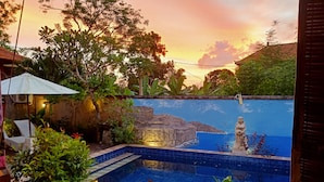 A swimming pool with a rock formation and a statue, surrounded by lush greenery and a sunset view.