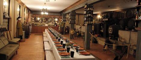 Una larga mesa de comedor dispuesta con platos, vasos y cubiertos, flanqueada por sofás y una zona de bar con botellas y barriles.